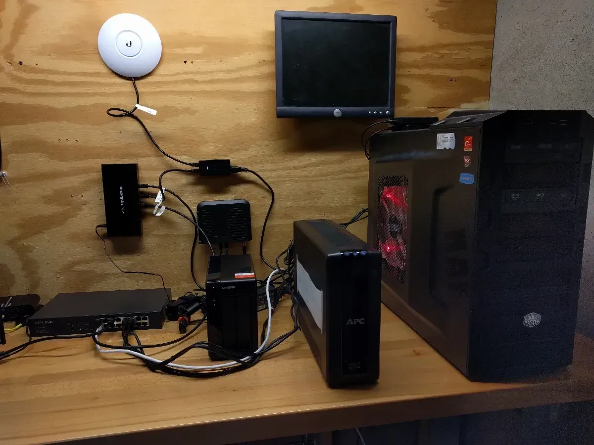 Early homelab workbench with small computers, a wall-mounted monitor, access point, and tower PC.