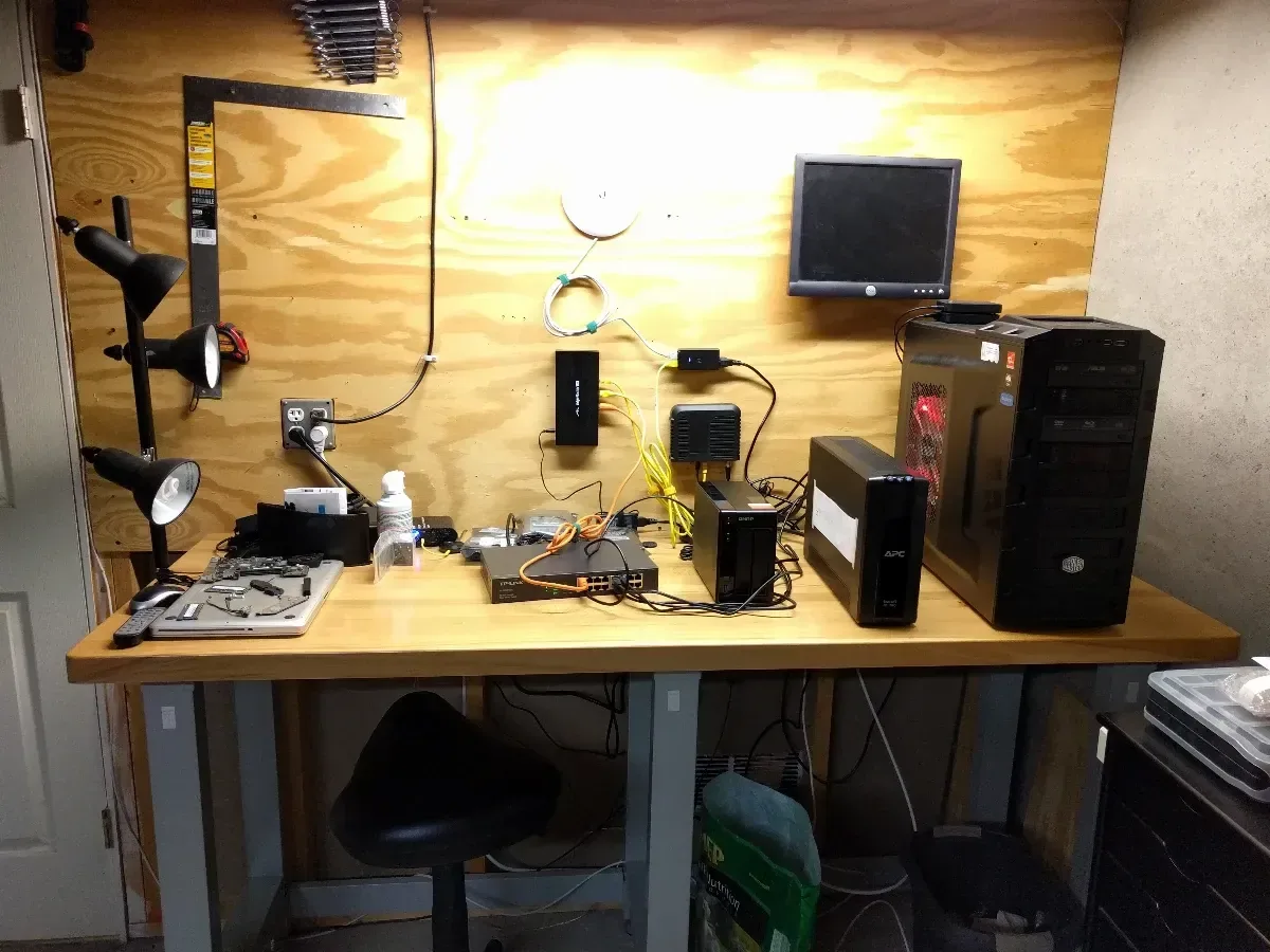 Wide view of the first homelab workbench beneath wall-mounted lighting and a monitor.