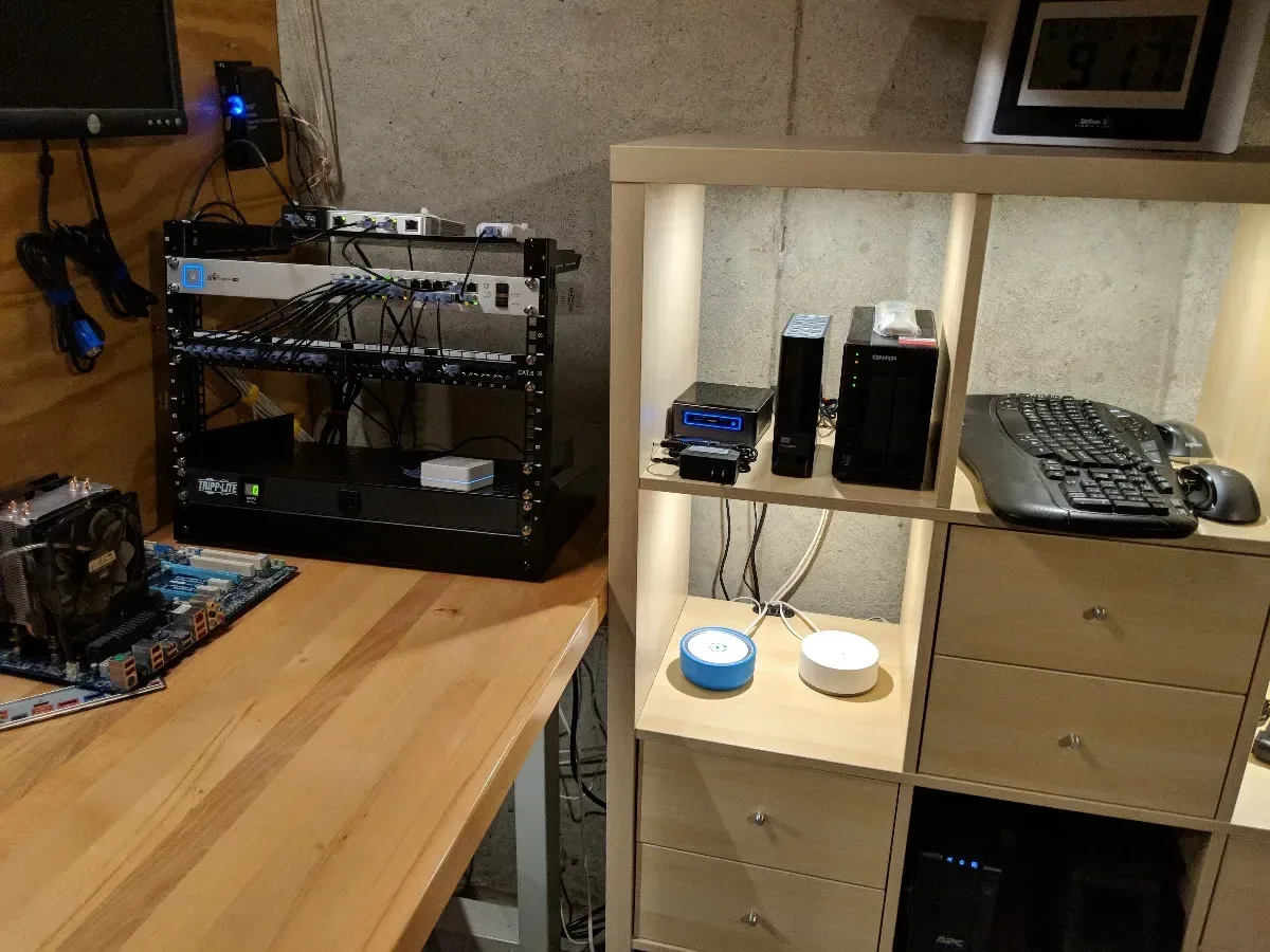 Open-frame network rack beside a storage shelf holding a tower server and small devices.