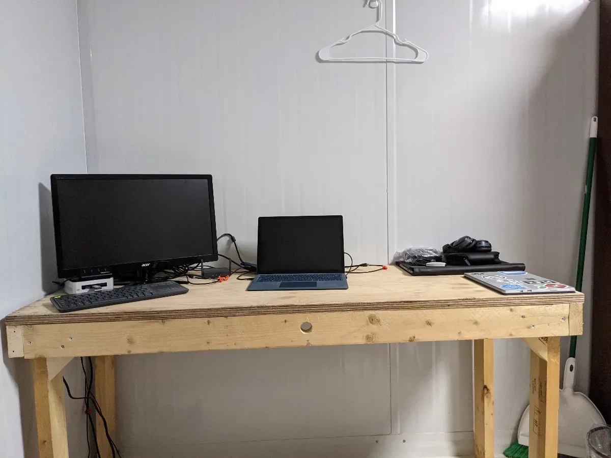 Simple wooden desk with a monitor, laptop, and compact homelab devices.