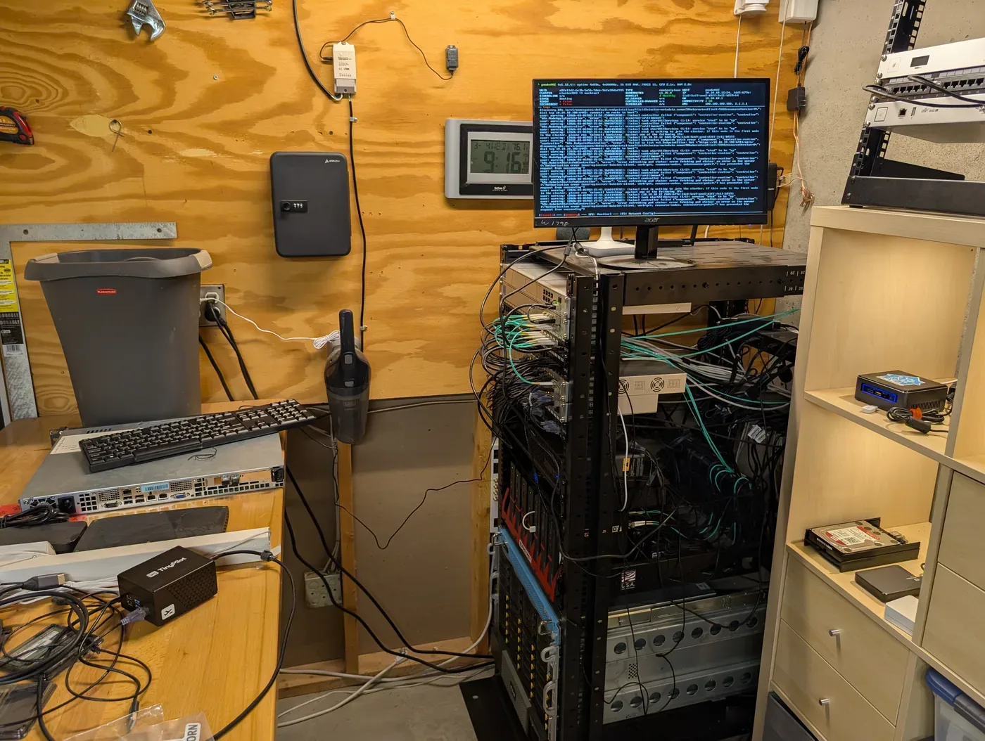 Wide view of the homelab workspace with a monitor, network rack, fiber cabling, and bench equipment.
